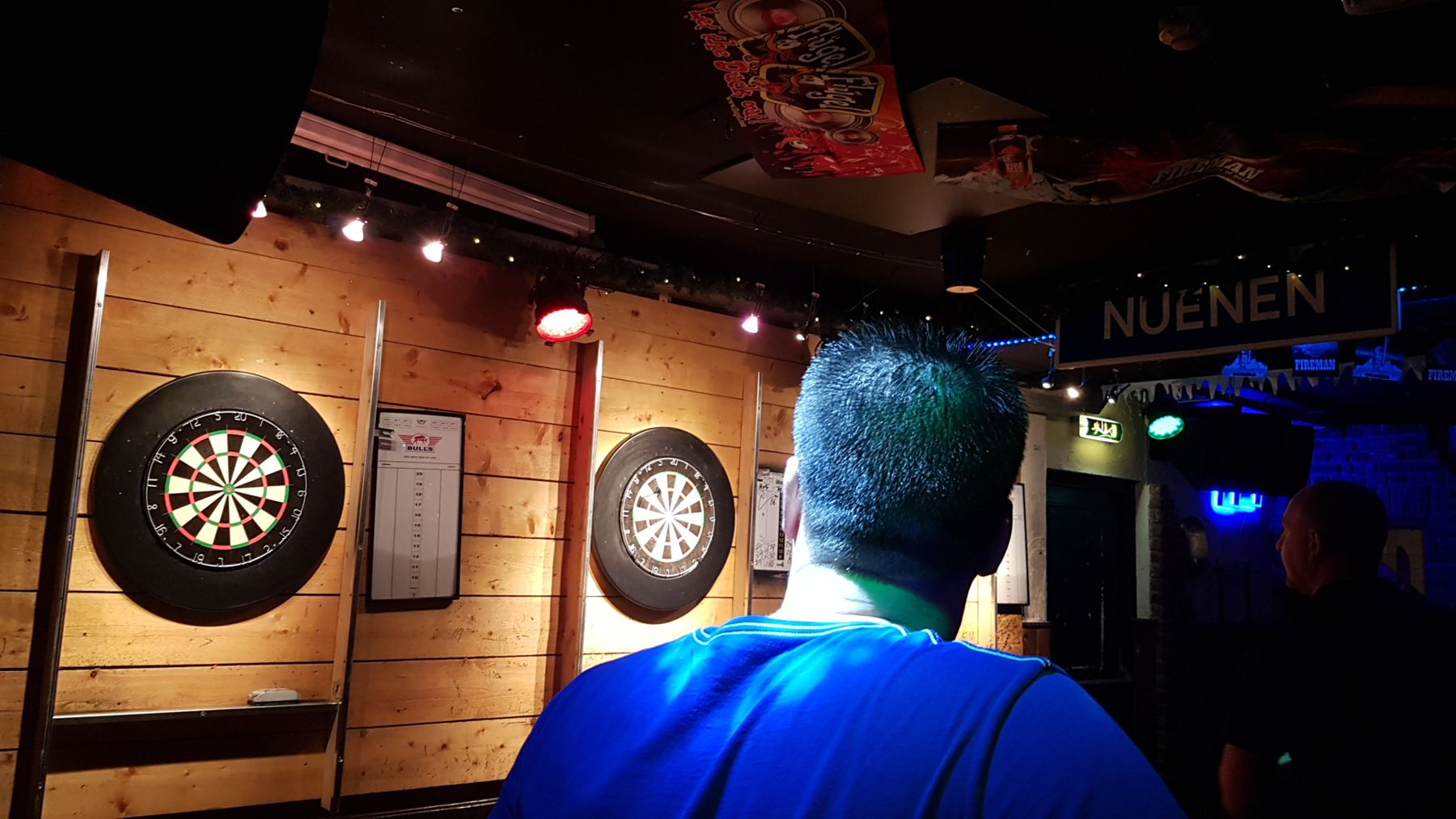 Twee dartborden aan een houten wand in ’t Hart van Son, een speler kijkt toe